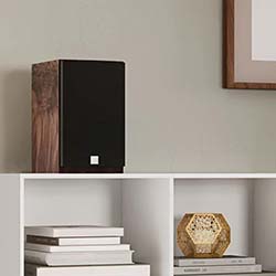 A black speaker with a wooden side sits on a white shelf. Nearby are stacked books and a geometric candle holder, creating a modern, minimalist decor vibe.
