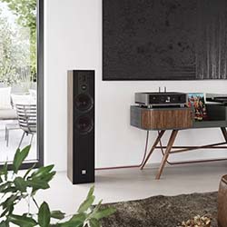 A modern living room with a tall black speaker, a wooden console holding a turntable, and plants. A glass door opens to a patio, creating a serene ambiance.