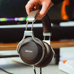 A hand holding brown Denon over-ear headphones in a cozy, tech-themed setting. Background includes a wooden table and blurred TV screen.