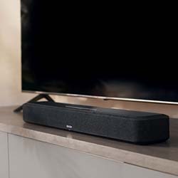 A sleek black soundbar sits atop a modern cabinet, positioned in front of a large flat-screen TV. The ambient setting conveys a minimalist style.