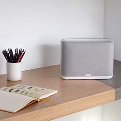 A minimalist desk setup with a modern gray speaker, a white cup holding black pencils, and an open sketchbook on a wooden surface, creating a calm atmosphere.