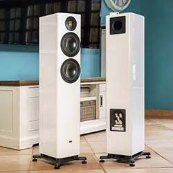 Two sleek white tower speakers stand on a tiled floor in a modern living room with light blue walls. The setting is elegant and high-tech.