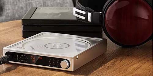 A sleek, metallic portable CD player with an open lid sits on a wooden table beside luxurious, maroon-cupped headphones, evoking a retro-modern vibe.