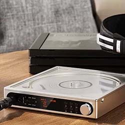 A sleek, metallic portable CD player with an open lid sits on a wooden table beside luxurious, maroon-cupped headphones, evoking a retro-modern vibe.