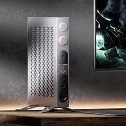 Silver, rectangular smart speaker with mesh front, dual dials, and a display screen sits on a wooden surface beside a glowing TV screen in a dim room.