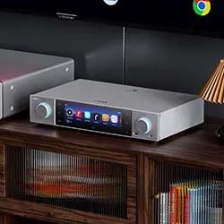 Silver audio amplifier with a digital display, flanked by two knobs, sits on a wooden shelf. The scene evokes a sleek, modern ambiance.