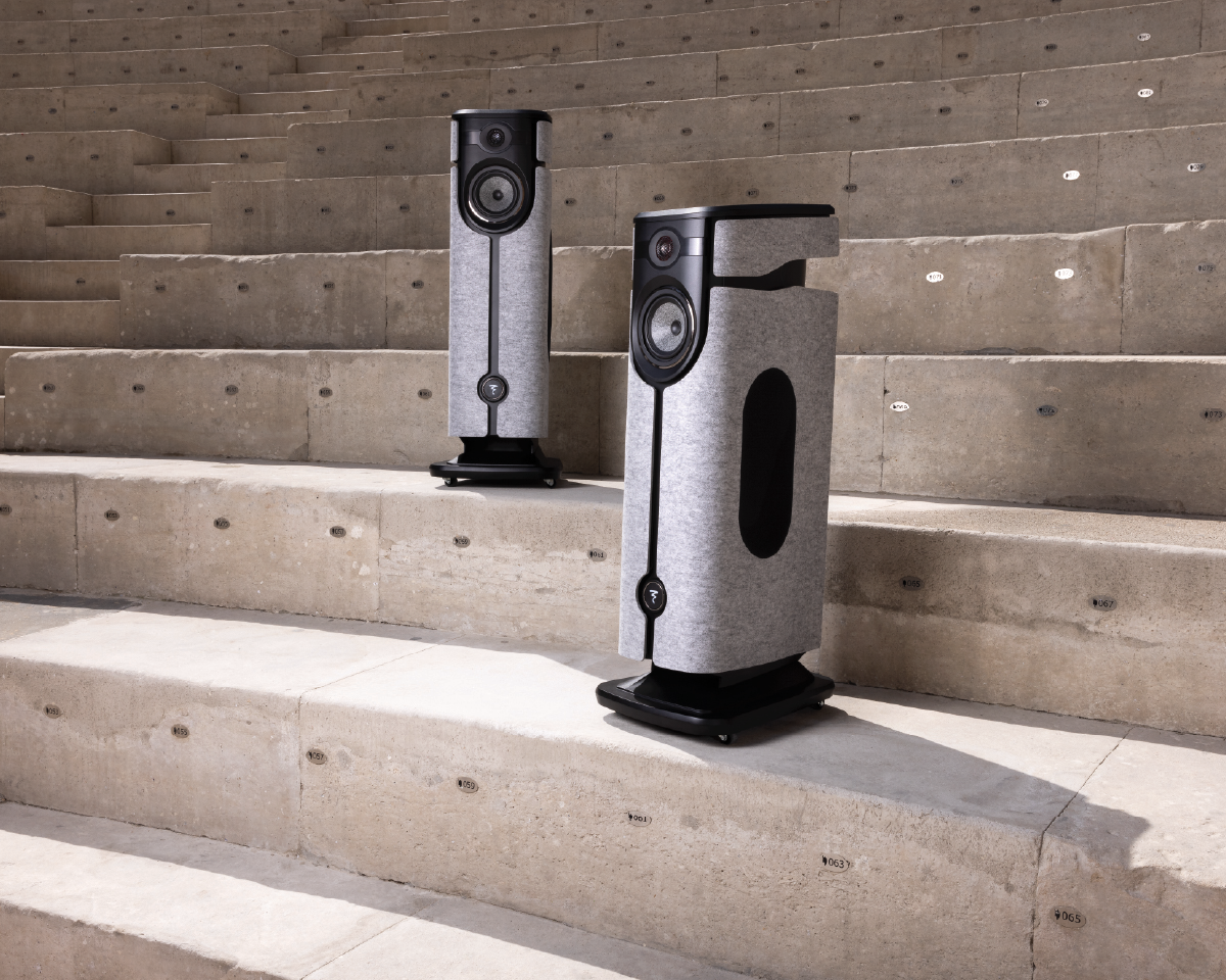 Two modern speakers with a sleek design stand on concrete steps. Both have a gray fabric covering and black accents, suggesting a minimalist aesthetic.