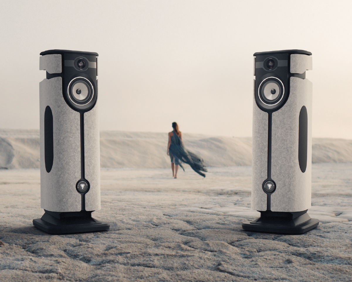 Two modern speakers stand prominently on a sandy background. In the distance, a person in a flowing dress walks away, adding a serene, surreal atmosphere.