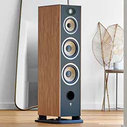 Tall wooden speaker with three circular drivers and a tweeter, set on a wooden floor next to a large mirror and decorative branches, conveying a modern and elegant vibe.