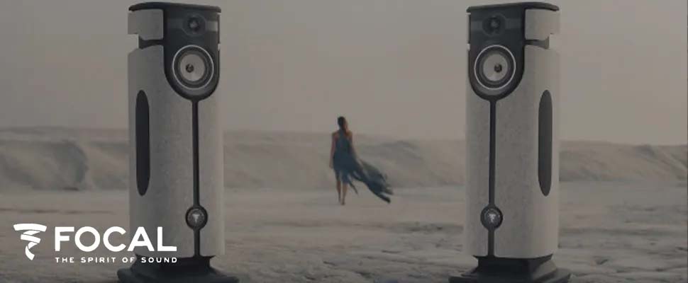 Two sleek speakers stand prominently in a vast desert landscape, with a lone person in a flowing dress walking away in the background. The scene conveys a sense of solitude and sophistication. Logo text reads "Focal: The Spirit of Sound" in the corner.