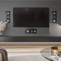 Modern living room with a wall-mounted flat-screen TV, flanked by sleek black speakers. A glass coffee table and neutral decor create a minimalist, cozy feel.