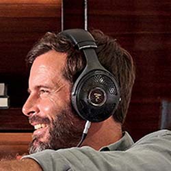 A smiling man with a beard enjoys music through large headphones, sitting comfortably against a wooden background, conveying relaxation and joy.