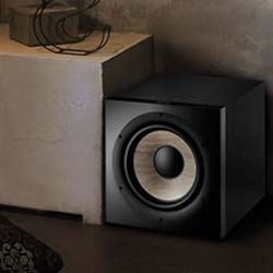A black subwoofer rests on a dark wood floor in a dimly lit room, next to a textured concrete wall and a light-colored table with coiled cables on top.