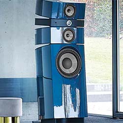Tall blue high-end speaker with multiple drivers stands in a modern, light-filled room by a window, conveying luxury and sophistication.