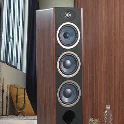 Tall brown floor-standing speaker with four circular drivers set against a wood-paneled wall, near a window. Modern, sleek design in a cozy room.