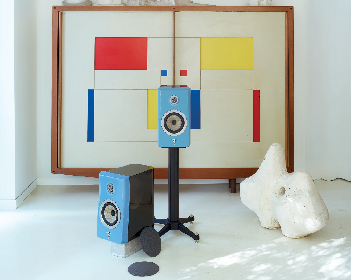 Blue speakers are displayed in front of a geometric abstract painting with red, yellow, and blue blocks. A white sculpture sits on the floor nearby, creating a modern, artistic atmosphere.