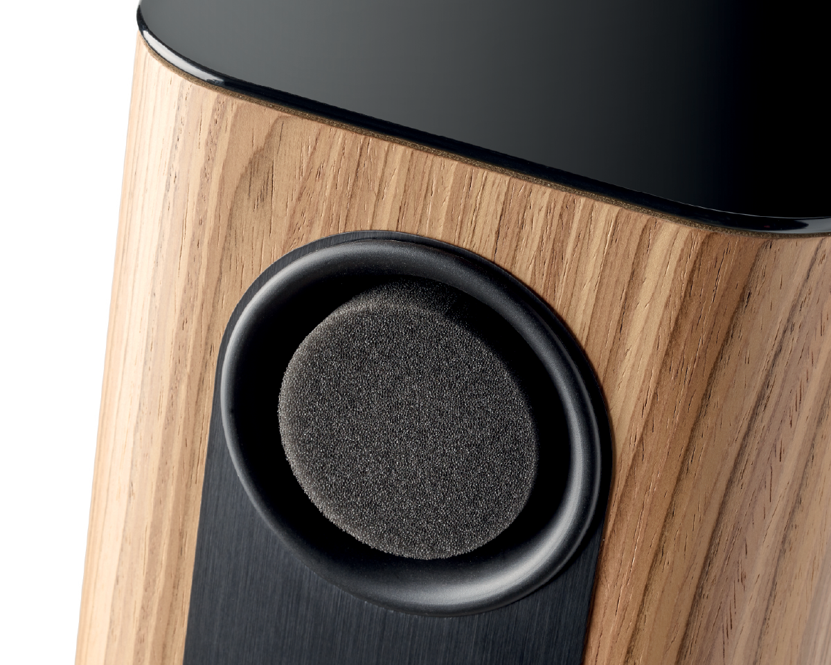 Close-up of a speaker with a wooden finish. The round, foam-covered speaker contrasts with the sleek, black and wood grain surface, conveying a modern design.