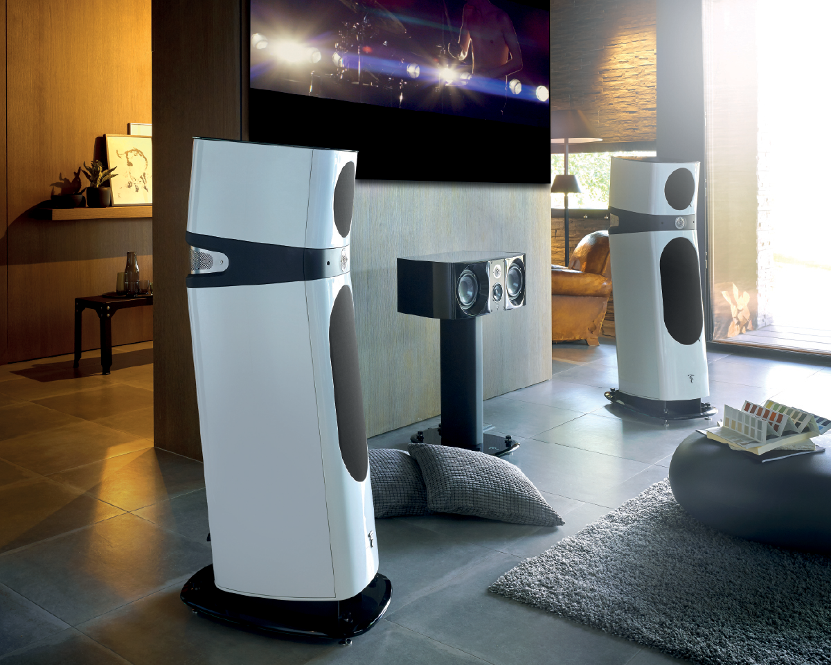 Modern home theater with sleek white floor-standing speakers and a central speaker on a stand. A large screen displays a dimly lit concert scene. The setting exudes a contemporary and cozy ambiance with soft lighting and plush gray carpets.