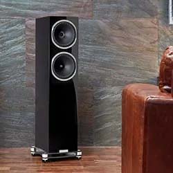 Tall black speaker with dual drivers on a wooden floor, placed against a stone-textured wall. A brown leather chair is partially visible, suggesting a cozy setting.