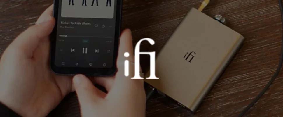 Hands hold a smartphone playing "Ticket to Ride" while connected to an iFi audio device on a wooden table. The atmosphere is cozy and focused on music enjoyment.