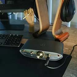 A sleek audio amplifier with a volume knob and headphone jack on a desk, next to a keyboard and wooden headphone stand, conveying a modern tech setup.