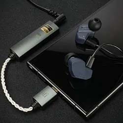 A sleek black smartphone, connected to blue in-ear headphones via a silver audio adaptor with a braided cable, lies on a dark surface, conveying a tech-savvy vibe.