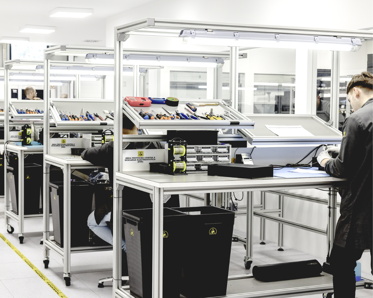 People work at individual stations in a clean, modern electronics assembly lab, with tools and equipment organized on shelves above each workstation.
