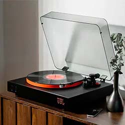 A modern turntable with a transparent lid plays a vinyl record on a wooden console. Next to it is a sleek black vase with green leaves.