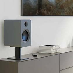 A sleek, gray speaker on a stand sits on a modern cabinet beside a stack of books. The scene conveys a minimalist and sophisticated tone.