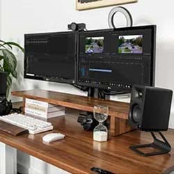 A tidy workspace featuring dual monitors displaying video editing software, a keyboard, mouse, speaker, and sand timer. Bright, organized, and tech-focused.