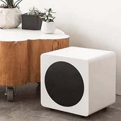 Modern white cube-shaped subwoofer with a black circular speaker sits on a concrete floor. Nearby is a wood table with small potted plants, creating a minimalist vibe.