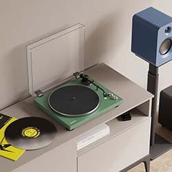 A green turntable sits on a modern wooden table with a clear cover open. A vinyl record is nearby. A tall, blue speaker stands in the background. The tone is retro and stylish.