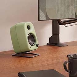 Modern desk setup with a green speaker on a stand, large monitor, black notebook, and mug. Warm tones convey a cozy, tech-savvy atmosphere.