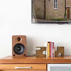 A modern living room with a wooden speaker, books, and abstract decor on a wooden console. A partially visible TV hangs above, showing a brick building.