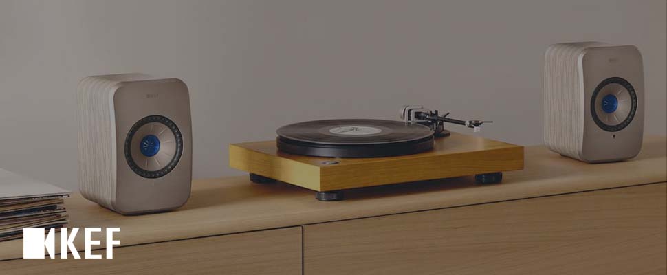 A sleek turntable with a wooden base is centered between two modern, compact speakers on a minimalist light wood cabinet. The tone is stylish and retro.
