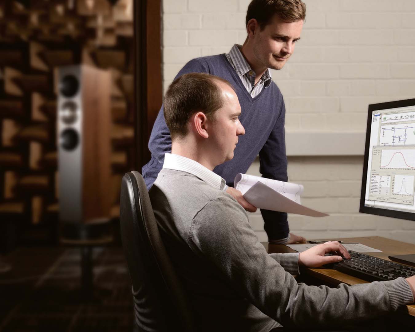 Two men in a soundproof room analyze data on a computer screen. One is seated, focused and clicking, while the other stands beside him, smiling and holding papers.