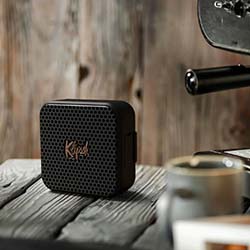 A small black portable speaker with a mesh front sits on a rustic wooden table beside a coffee cup, conveying a relaxed, cozy ambiance.