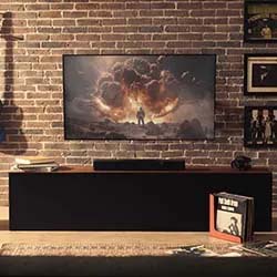 A TV mounted on a brick wall displays a dramatic scene with explosions. A guitar and framed photos hang nearby, creating a cozy, artistic vibe.