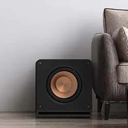 Black subwoofer with a copper speaker cone sits on a wooden floor next to a striped gray sofa, against a plain white wall, creating a sleek, modern vibe.