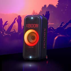 Portable speaker with vibrant, glowing orange and red lights labeled "XBOOM" in front of a lively concert crowd with raised hands against a purple-lit sky.