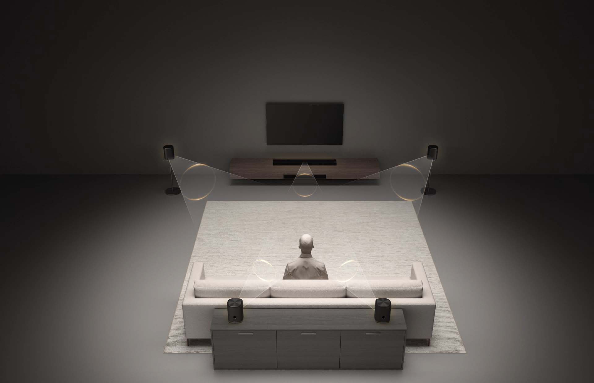 A person sits on a gray couch in a dimly lit room, facing a TV. Surround speakers create an immersive sound setup, enhancing a home theater experience.