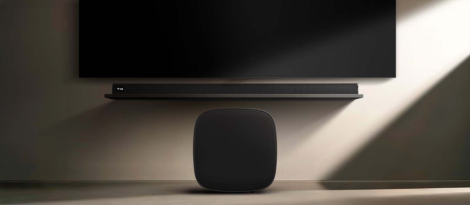 A sleek, modern entertainment setup features a wall-mounted TV, a slim soundbar beneath, and a compact black subwoofer on the floor, under soft lighting.