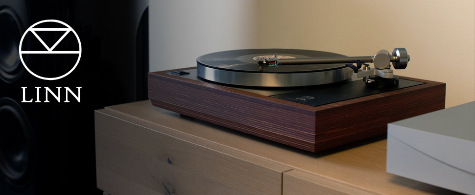 A sleek Linn record player with a wooden base sits on a wooden surface, exuding a classic, refined aesthetic. A speaker is visible in the dimly lit background.