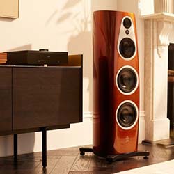 Tall, sleek brown speaker with multiple drivers sits beside a dark wood cabinet in a stylish living room, creating a warm, modern atmosphere.
