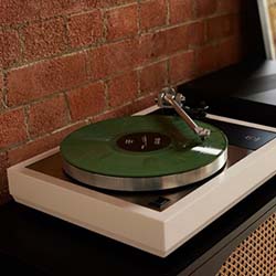 A green vinyl record sits on a modern white turntable next to a brick wall, creating a nostalgic yet contemporary vibe in a cozy room.