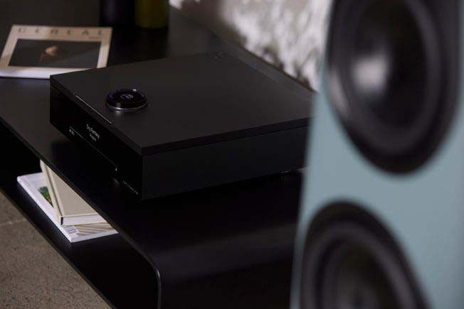 A sleek black audio receiver with a round control knob is on a dark shelf. Nearby, a large speaker and open magazine add an elegant, modern vibe.
