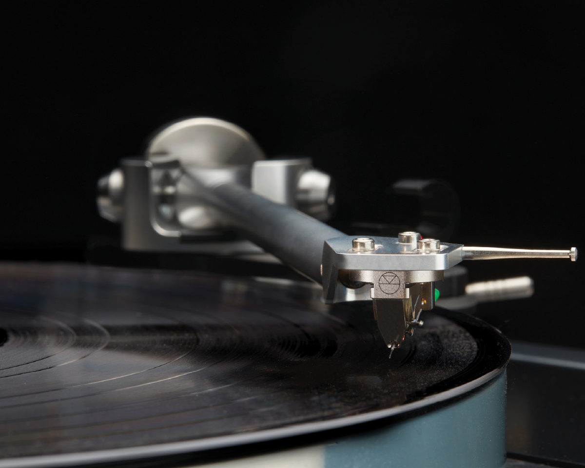 Close-up of a turntable needle touching a black vinyl record against a dark background. The metal arm is focused and detailed, evoking a vintage feel.