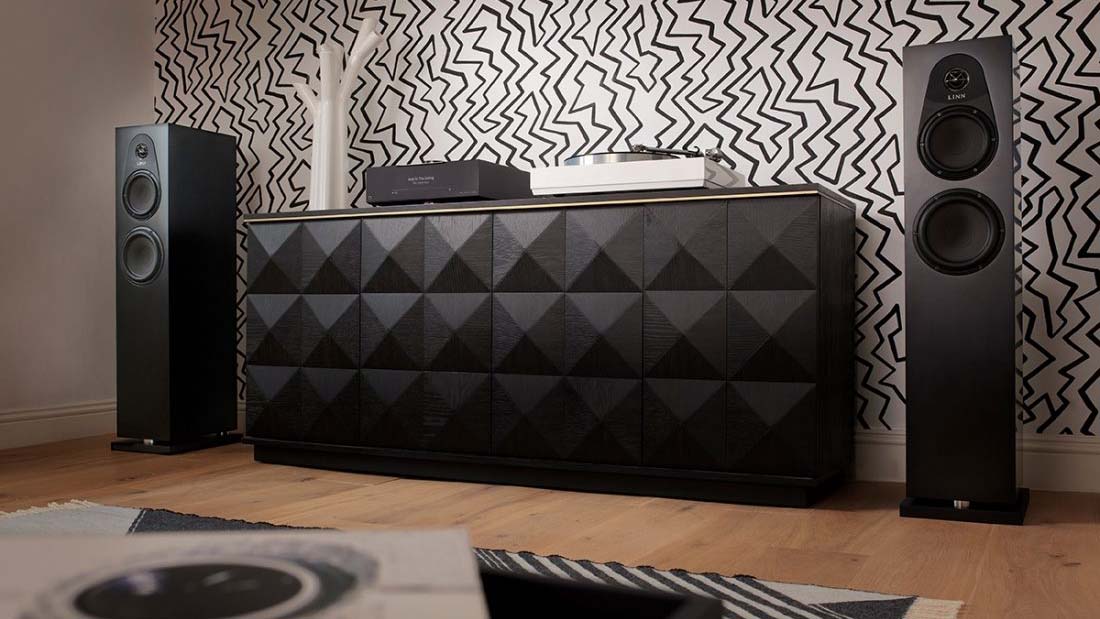 Modern audio setup with black speakers flanking a geometric cabinet, turntable, and wireless equipment. Zigzag pattern wallpaper adds a lively backdrop.