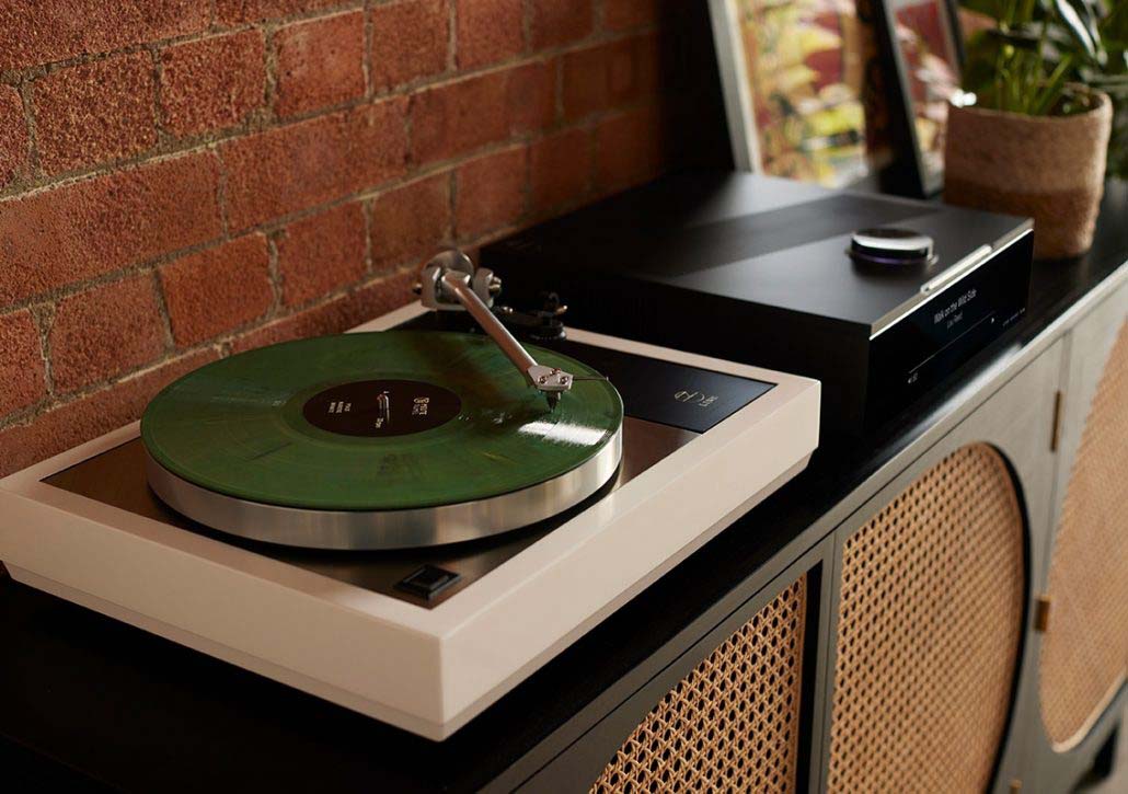 A stylish white turntable with a green vinyl record sits on a cabinet against a brick wall. Nearby, a sleek black audio device adds a modern touch.