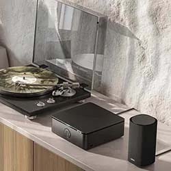 A modern turntable with a marbled vinyl record under a clear dust cover, next to a sleek black amplifier and speaker, set on a wooden shelf.
